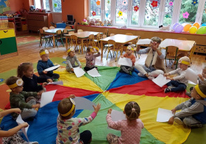 Nauczycielka i przedszkolaki podnoszą do góry białe kartki tworząc wiatr.