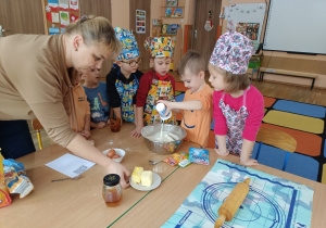Dzieci ubrane w stroje kucharskie wsypują składniki do miski.