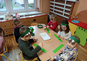 Dzieci siedzą przy stolikach i kleją opaski dinozaurów