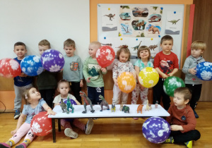 Dzieci z grupy Gwiazdeczki przy tablicy demonstracyjnej z balonami i figurkami dinozaurów.