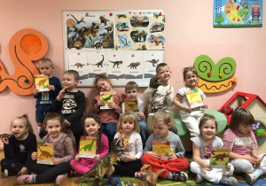 Przedszkolaki z gr. III na tle tablicy demonstracyjnej pokazują obrazki dinozaurów.