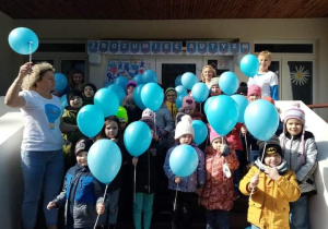 Dzieci z najstarszych grup stoją przed wejściem do przedszkola z niebieskimi balonami.