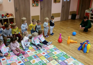 Dzieci siedzą, nauczycielka demonstruje sposób gry w kręgle.
