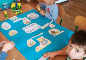 Dzieci przyklejają obrazki do dużego niebieskiego kartonu tworząc plakat "Prawa dziecka"