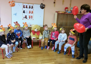 Dzieci z nauczycielką siedzą na krzesełkach w kręgu, nauczycielka stoi obok trzyma w ręku balony.