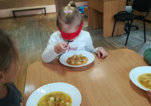 Dziewczynka z zasłoniętymi oczami czerwoną chustką rozpoznaje zupę po smaku.