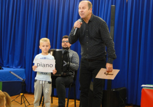 Pan z akordeonem, pan z mikrofonem i chłopiec trzymający napis „piano”.