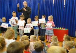 Pan z mikrofonem i grupa dzieci trzymająca kartki z nazwami solmizacyjnymi.