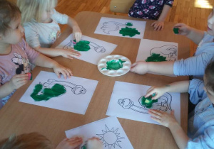 Dzieci stemplują gąbką i farbą w kolorze zielonym rysunek dinozaura.