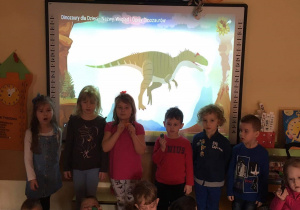 Zdjęcie dzieci z grupy IV z figurkami dinozaurów na tle tablicy multimedialnej.
