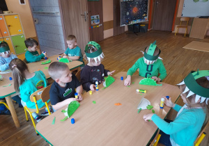 Dzieci siedzą przy stoliku i wykonują dinozaura z papierowego talerzyka pomalowanego na zielono.