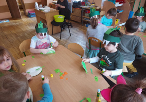 Dzieci siedzą przy stoliku i wykonują dinozaura z papierowego talerzyka pomalowanego na zielono.