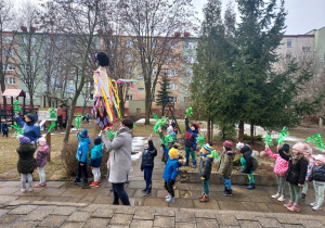 Dzieci wyszły do ogrodu z zielonymi miotełkami w ręku, pani trzyma Marzannę - przeganiają zimę