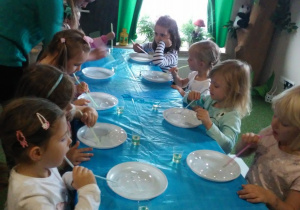 Dzieci siedzą przy stoliku, przed nimi są plastikowe talerze z cieczą w którą dmuchają przez słomkę.