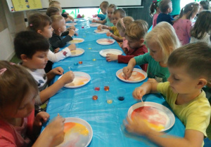 Dzieci siedzą przy stoliku, przed nimi są plastikowe talerze na których barwią ciecz.