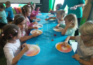 Dzieci siedzą przy stoliku, przed nimi są plastikowe talerze na których barwią ciecz., przed nimi są plastikowe talerze na których barwią ciecz.