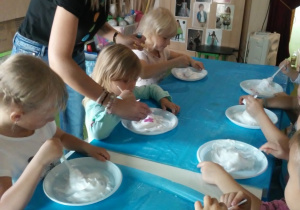 Dzieci siedzą przy stoliku, przed nimi są plastikowe talerze na których mają białą substancję.
