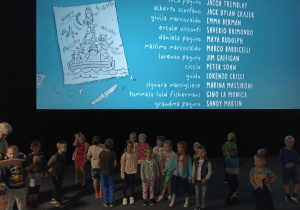 Dzieci stoją pod dużym ekranem na którym są napisy końcowe filmu.