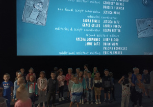 Dzieci stoją pod dużym ekranem na którym są napisy końcowe filmu.