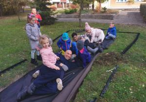 Pan Rafał, pani dyrektor i dzieci leżą na płachcie materiału w ogrodzie