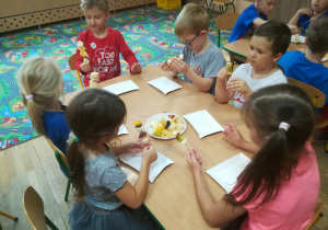 Dzieci siedzą przy stoliku i nadziewają pokrojone owoce na długie wykałaczki.