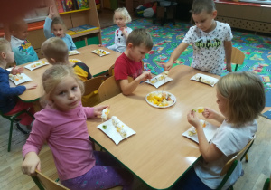Dzieci siedzą przy stoliku i nadziewają pokrojone owoce na długie wykałaczki.
