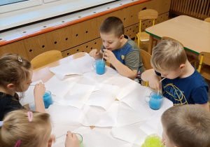 Dzieci siedzą przy stolikach i dmuchają przez słomki do szklanek z kolorową cieczą