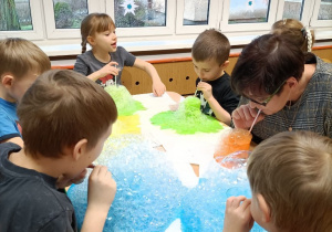 Dzieci siedzą przy stolikach i dmuchają przez słomki do szklanek z kolorową cieczą tworząc pianę
