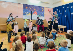 Muzycy grają i śpiewają dla przedszkolaków, na środku stoją trzy pary przedszkolaków w kapeluszach