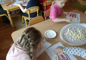 Dzieci siedzą przy stolikach i tworzą pracę plastyczną z popcornem