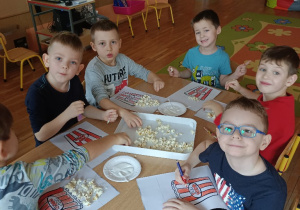 Dzieci siedzą przy stolikach i tworzą pracę plastyczną z popcornem