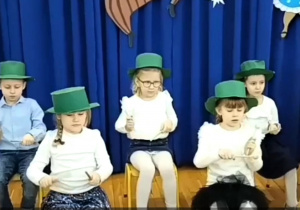 Dzieci siedzą na krzesełkach w zielonych kapeluszach tworząc orkiestrę
