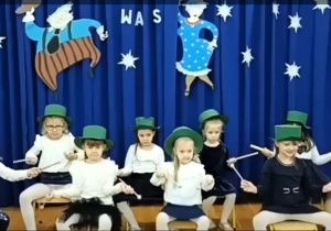 Dzieci siedzą na krzesełkach w zielonych kapeluszach tworząc tańczącą orkiestrę