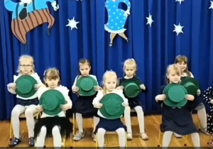 Dzieci siedzą na krzesełkach w zielonych kapeluszach tworząc orkiestrę