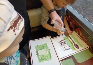Dzieci z pomocą osoby dorosłej wypełniają czekoladą silikonowe foremki