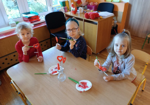 Trzy dziewczynki siedzą przy stoliku i malują czerwoną farbą styropianowe serduszka