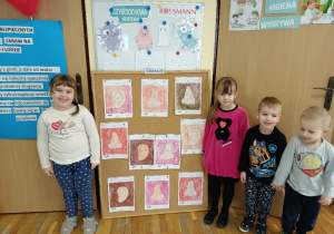 Dzieci stoją przy tablicy na której są przyczepione pokolorowane obrazki