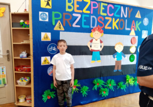 Mateusz stoi na tle dekoracji „Bezpieczny Przedszkolak”
