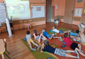 Dzieci leżąc na dywanie oglądają bajkę o pieskach na dużym ekranie