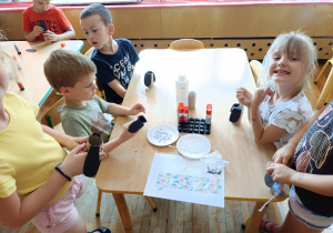 Dzieci siedzą przy stoliku i wykonują pieska z rolki po papierze toaletowym.