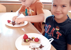 Chłopcy dekorują herbatniki czekoladą, truskawkami i posypkami.