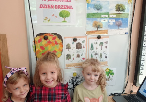 Trzy dziewczynki stoją na tle tablicy na której sa informacje o drzewach
