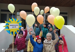 Dzieci z grupy „Słoneczka” z balonami i transparentem stoją przed przedszkolem.