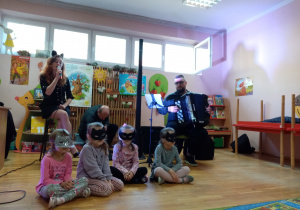 Wokalistka śpiewa piosenkę, a dziewczynki w maskach kotów siedzą na scenie.