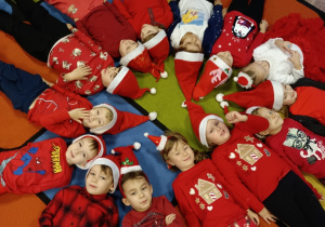 Dzieci leżą na dywanie na plecach w kółeczku, w czerwonych czapkach.