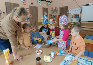 Dzieci ubrane w stroje kucharskie wsypują składniki do miski.