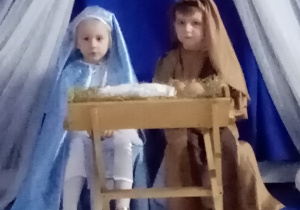 Dzieci odgrywające rolę Maryi i Józefa siedzą przy żłóbku.