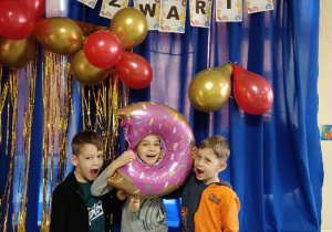 Pamiątkowe zdjęcie trójki dzieci z dmuchanym balonem w kształcie pączka na tle napisu Tłusty Czwartek