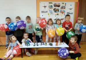 Dzieci z grupy Gwiazdeczki przy tablicy demonstracyjnej z balonami i figurkami dinozaurów.