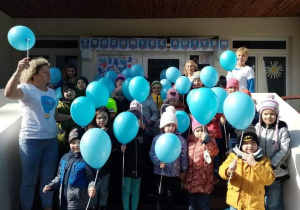 Dzieci z najstarszych grup stoją przed wejściem do przedszkola z niebieskimi balonami.